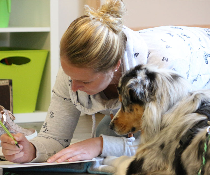 Eine Frau schreibt, lächelt und ein Hund schaut zu. Beide konzentriert; Zimmereinrichtung im Hintergrund.